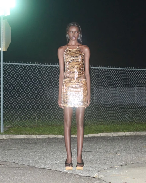 Elegant adult woman in a shimmering halter dress and heels poses in an outdoor nighttime fashion shot against a chain-link fence.