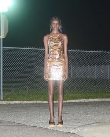 Elegant adult woman in a shimmering halter dress and heels poses in an outdoor nighttime fashion shot against a chain-link fence.