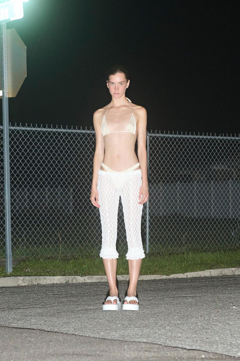 A slender woman in a light bikini and crochet pants poses at night in front of a chain-link fence beneath the street light.