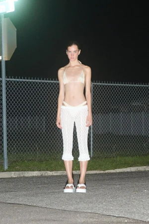 A slender woman in a light bikini and crochet pants poses at night in front of a chain-link fence beneath the street light.