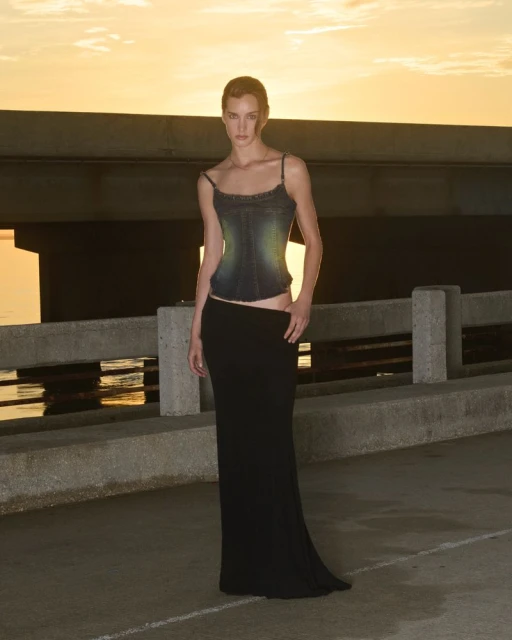 Fashionable adult female model in a two-piece outfit posing against a sunset backdrop with water, sky, and architecture.