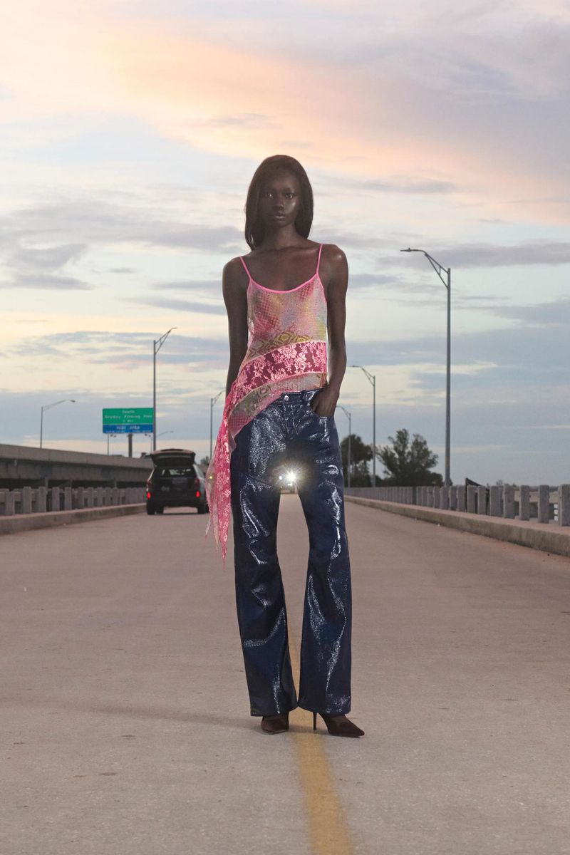 Fashionable Black woman poses on an empty road at dusk, wearing a sequined top and flared pants, with a car in the background.
