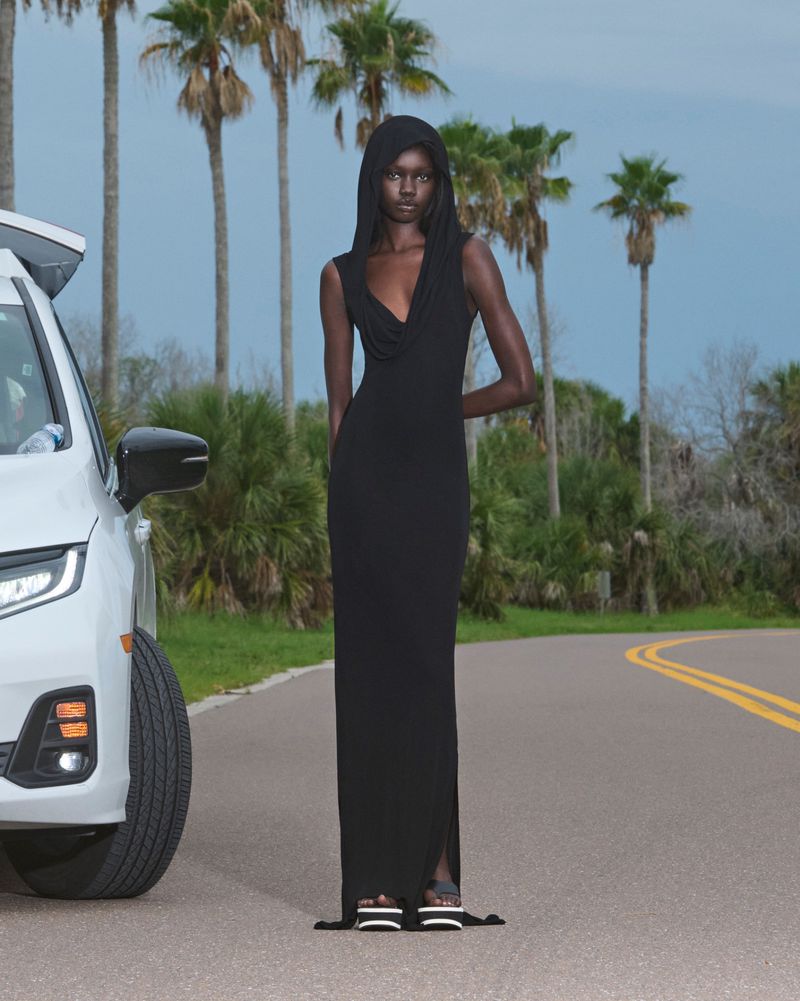 Woman in black dress poses on asphalt road next to parked white car with palm trees in background under blue sky, fashion editorial.