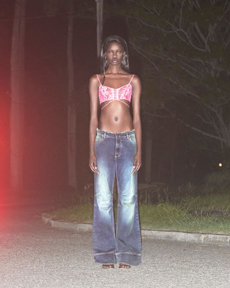 Fashionable woman in a pink bra and wide-leg jeans poses on a paved road at night, with trees and dimly lit surroundings.