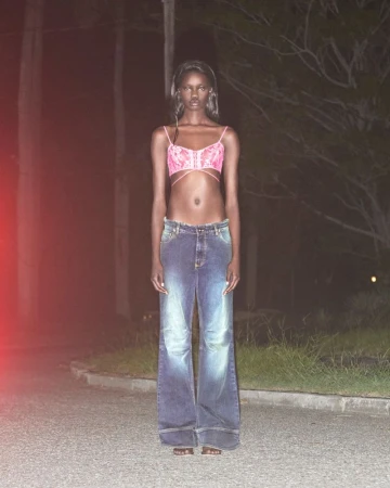 Fashionable woman in a pink bra and wide-leg jeans poses on a paved road at night, with trees and dimly lit surroundings.