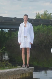 Model standing on a concrete ledge by the water wearing a white shirt and matching shorts for PRISCAVera.