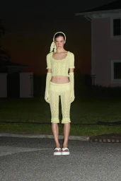 Model standing outdoors at night wearing a matching yellow knit top, shorts, headband, and gloves with white sandals.