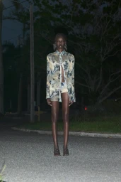 Model standing outdoors at night on a dark paved road wearing a patterned button-down shirt and shorts under direct flash lighting