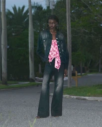Model standing on a paved street at dusk wearing a dark denim jacket and flared jeans over a pink patterned top