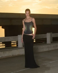 Model standing outdoors near a concrete bridge at sunset wearing a dark top and long skirt for PRISCAVera S/S 26