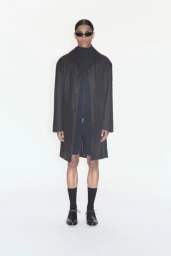 Full-length portrait of a male model wearing dark sunglasses, an oversized coat, zip-up top, shorts, and black shoes against a white background.