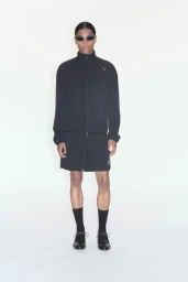Model wearing a black zip-up track jacket and matching shorts with dark sunglasses against a plain white background
