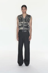 Model wearing a camouflage knit vest with bold lettering and black trousers against a plain white studio background
