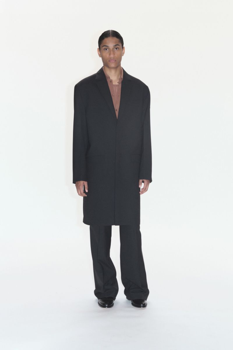 Full-length portrait of a slender adult man in a dark overcoat and pants against a bright white backdrop in a fashion studio.