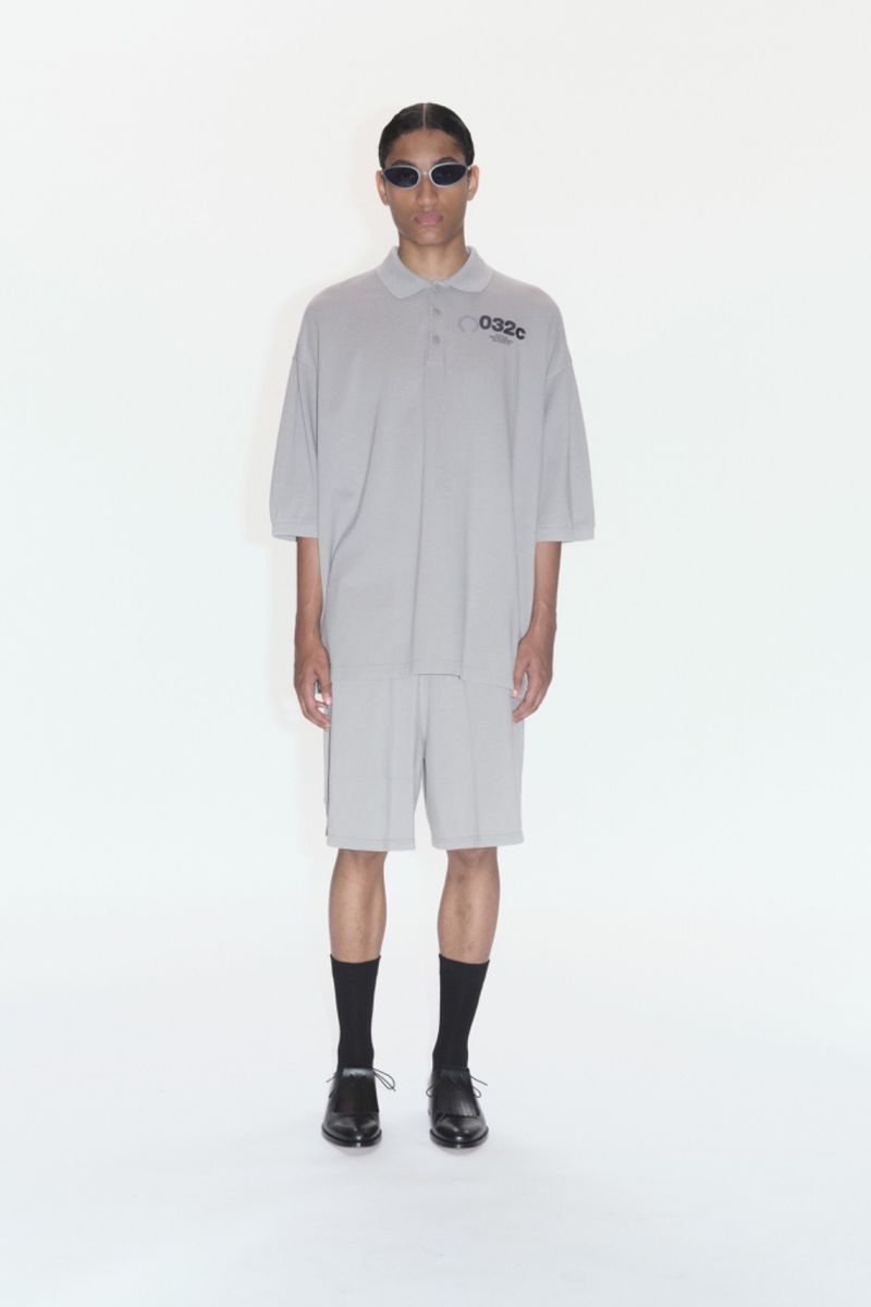 Male model poses in grey polo shirt and shorts with black socks and shoes against a white background, wearing sunglasses.