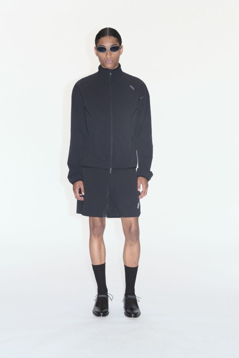A full-length shot of a male model in a black tracksuit with a jacket and shorts, wearing black shoes, socks, and futuristic sunglasses.
