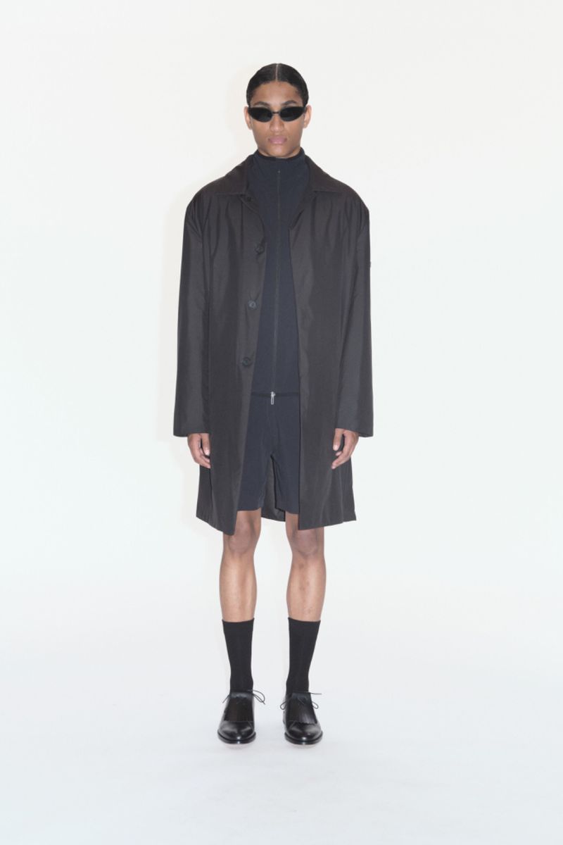 Studio shot of a person wearing a dark coat, shorts, socks, and shoes, styled with sunglasses, set against a plain white backdrop.