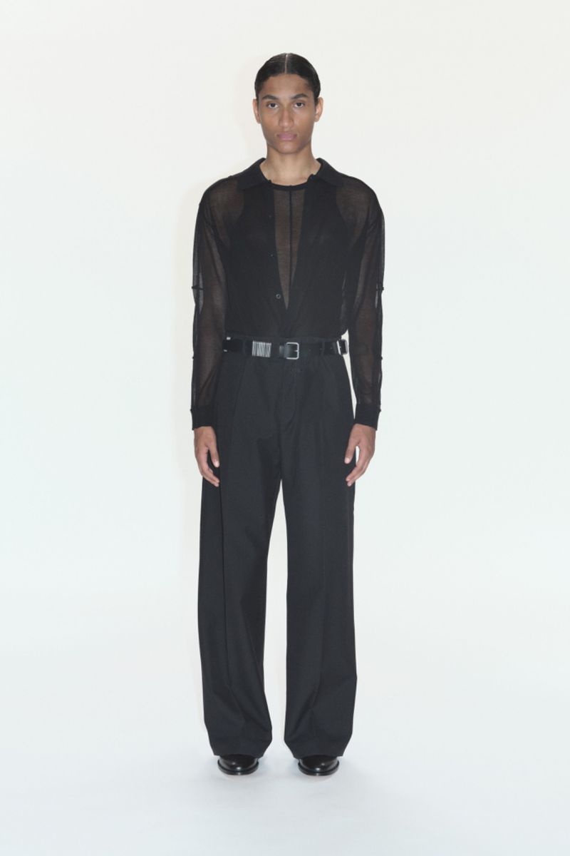 Full-length studio shot of a slender adult male posing, wearing a black mesh top, wide-leg black pants, and a silver belt against a white backdrop.