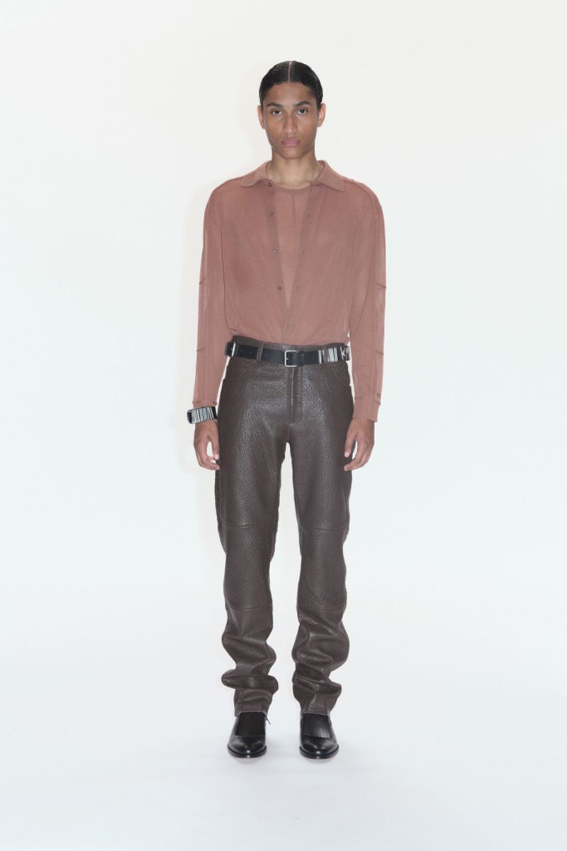 A solo male model stands in a photo studio wearing a tan shirt, brown leather pants, a black belt, and black leather shoes.