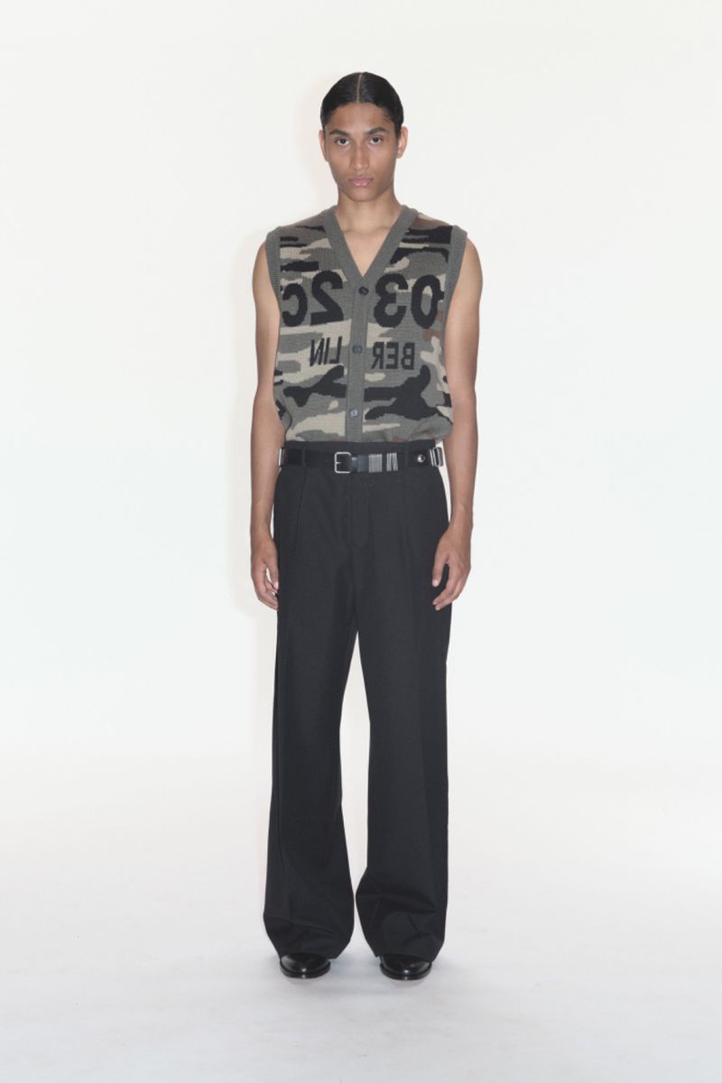 Male fashion model wearing a camouflage print sweater vest with black wide-leg pants standing in a white photo studio backdrop.
