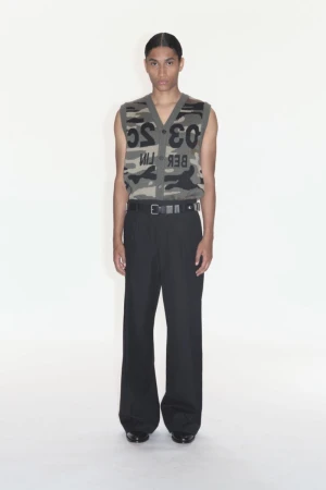 Male fashion model wearing a camouflage print sweater vest with black wide-leg pants standing in a white photo studio backdrop.