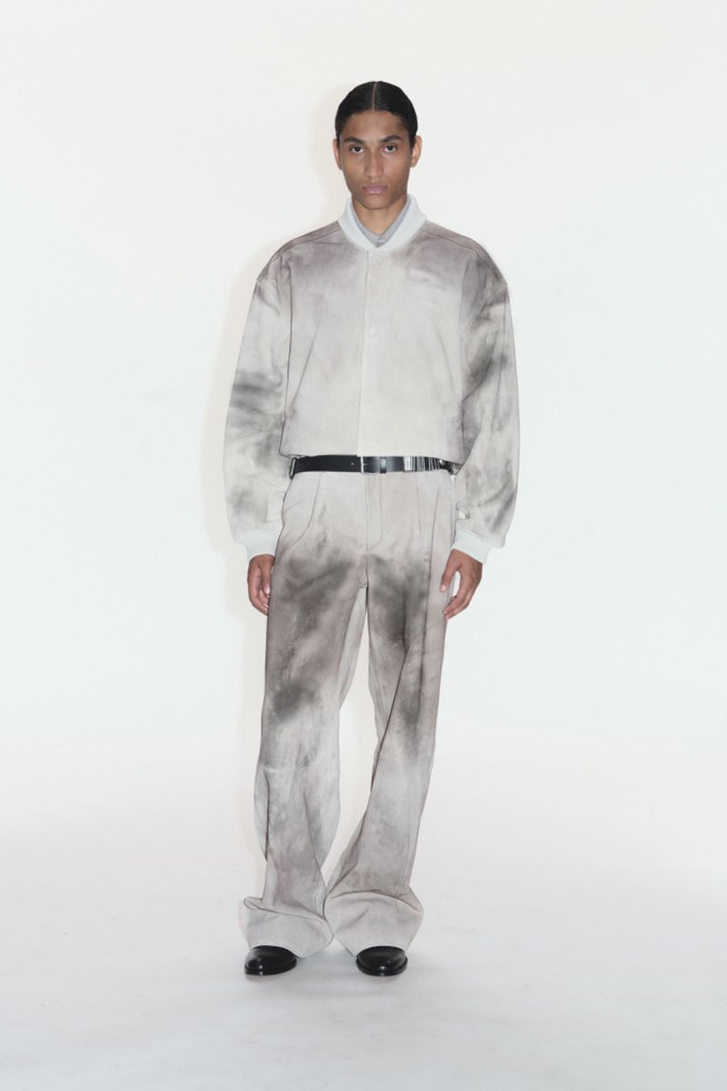 A male model poses in a studio wearing a light beige jacket and matching pants with dark grey abstract dye patterns and black leather dress shoes.