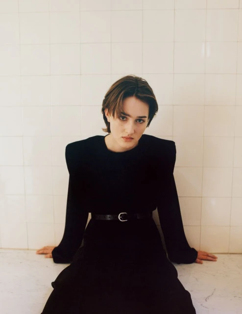 Adult female in a black dress with shoulder pads and a belt sits with hands on a white surface, in an indoor setting against tile.