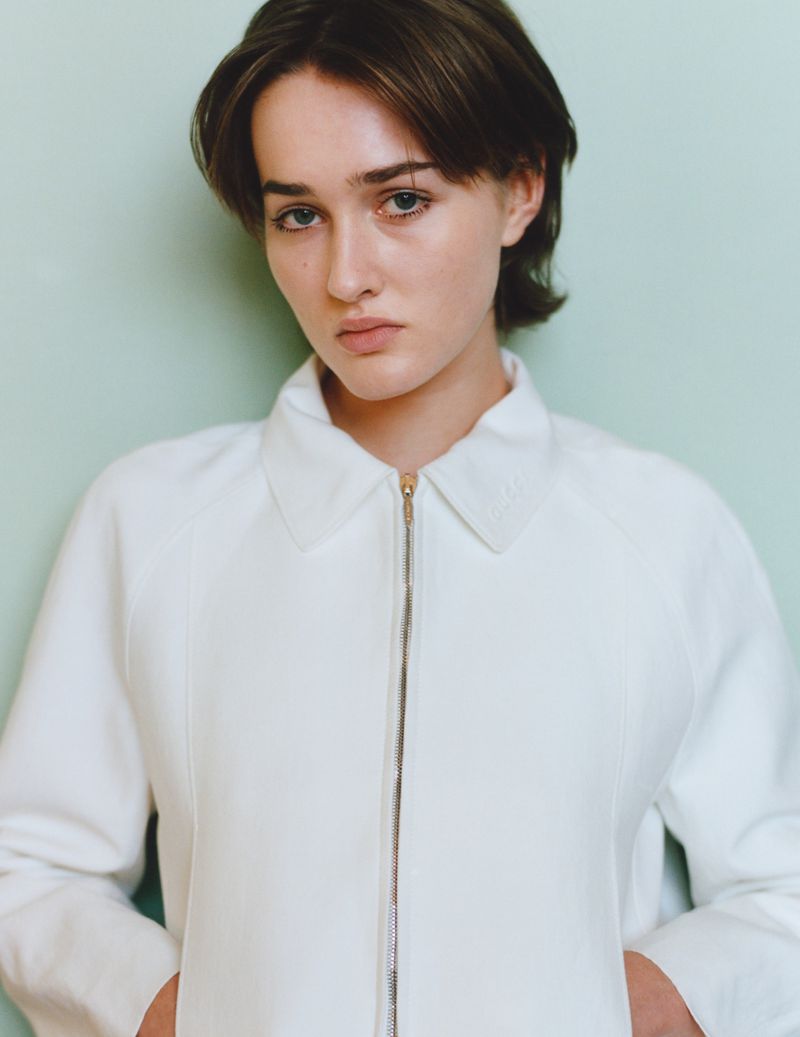 Close-up portrait of a young woman with short brown hair wearing a white zip-up jacket against a light green wall background.