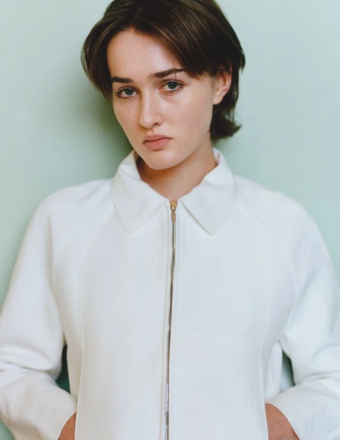 Close-up portrait of a young woman with short brown hair wearing a white zip-up jacket against a light green wall background.