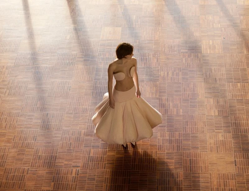 Aerial view of a female fashion model in a pale-pink strapless dress, standing on a brown parquet floor with sunlight and shadow.