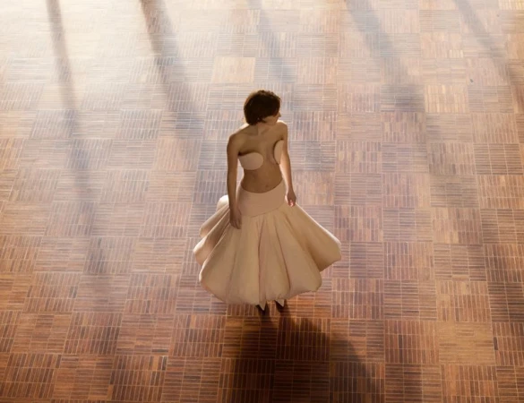 Aerial view of a female fashion model in a pale-pink strapless dress, standing on a brown parquet floor with sunlight and shadow.