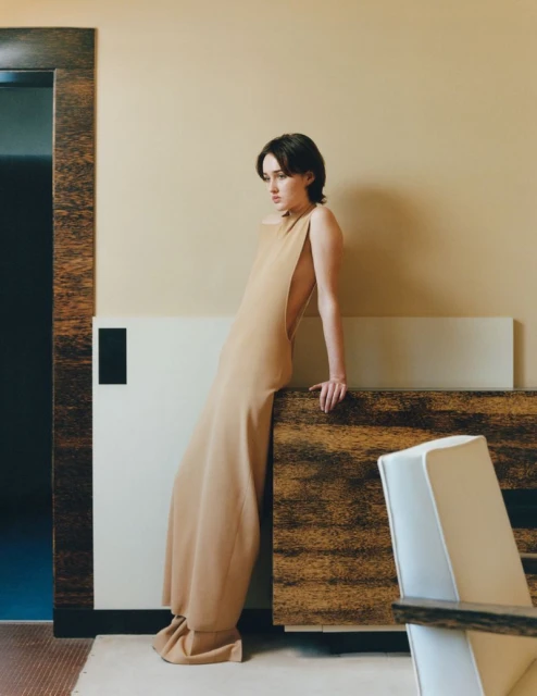Adult female model with short brunette hair leans against a textured wall in a long beige gown near a white modern chair indoors.