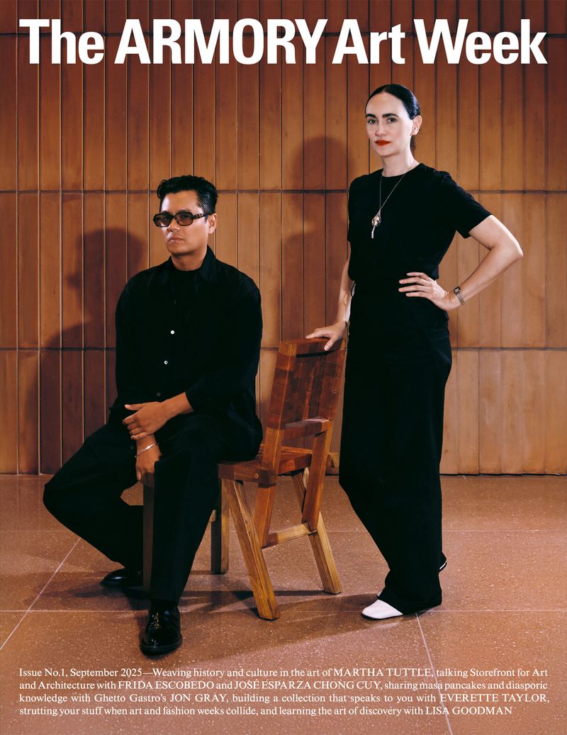 Magazine cover featuring a man seated on a wooden chair beside a standing woman with dark hair and wearing a black dress indoors.