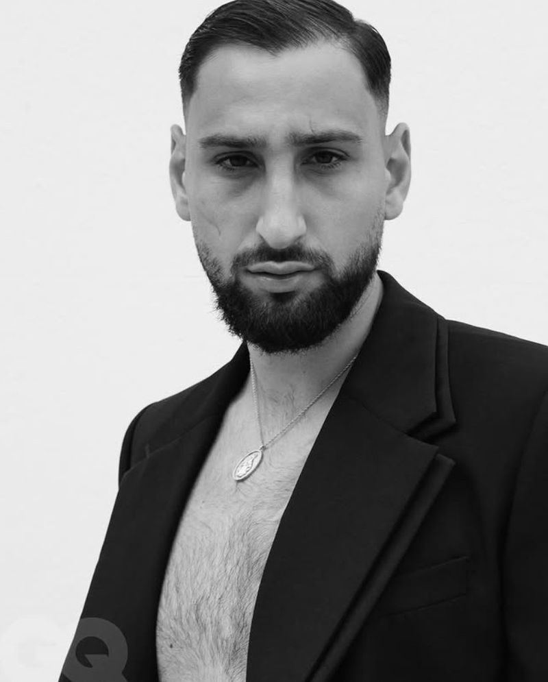 Black and white portrait of a stylish adult male with a beard wearing an open suit jacket and a necklace against a plain white background.