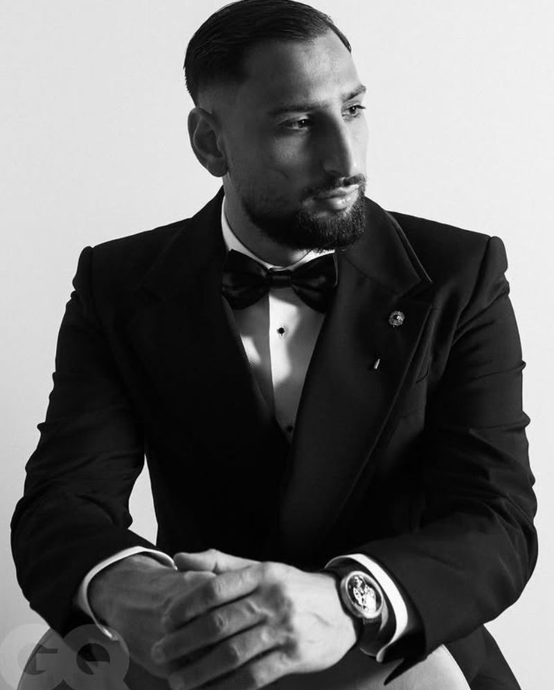 Monochrome portrait of a bearded man in a tuxedo with a bowtie, posing in a photo studio, showcasing formal wear and an elegant wristwatch.