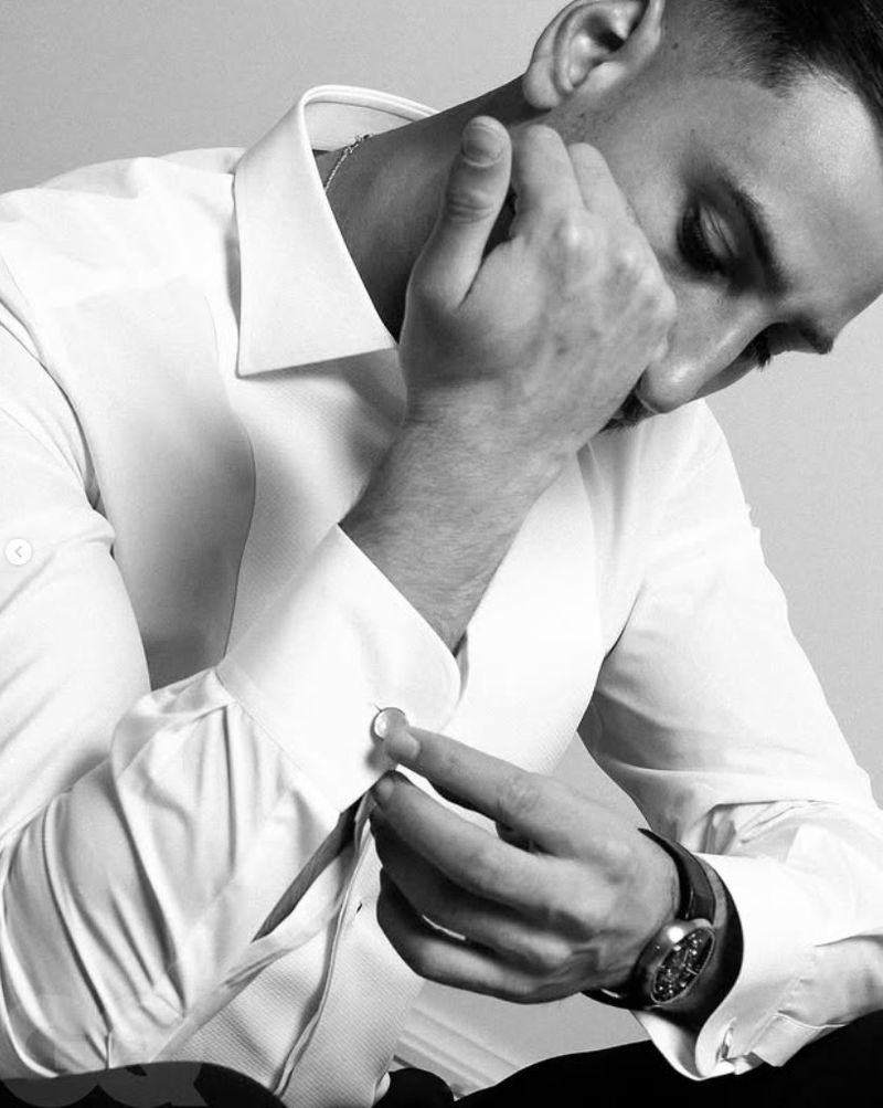 Monochrome close-up of a man in a white dress shirt, adjusting his cufflink with a focused expression, set against a neutral background.