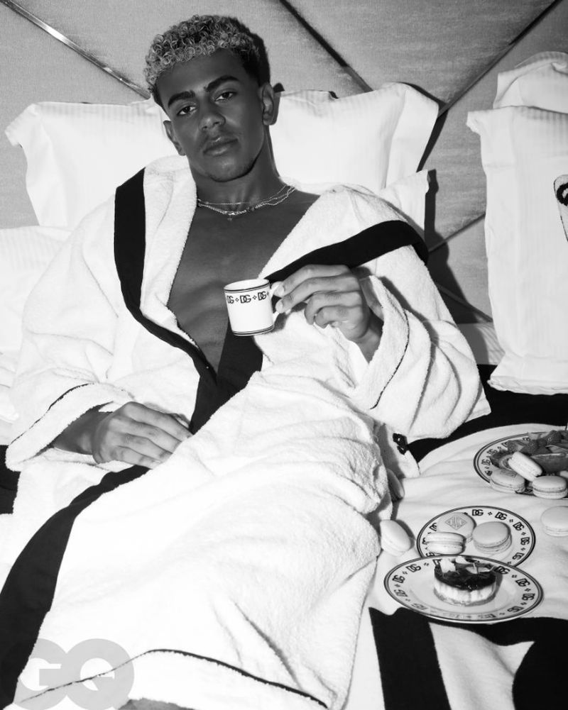 Black and white portrait of a man in a robe reclining in bed holding a teacup with pastries on plates on the bed.