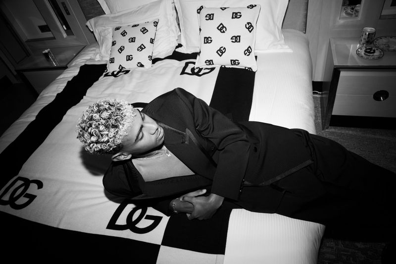 Monochrome image of a person with light curly hair, wearing a dark suit and lying on a Dolce & Gabbana branded bed in a dimly lit room.