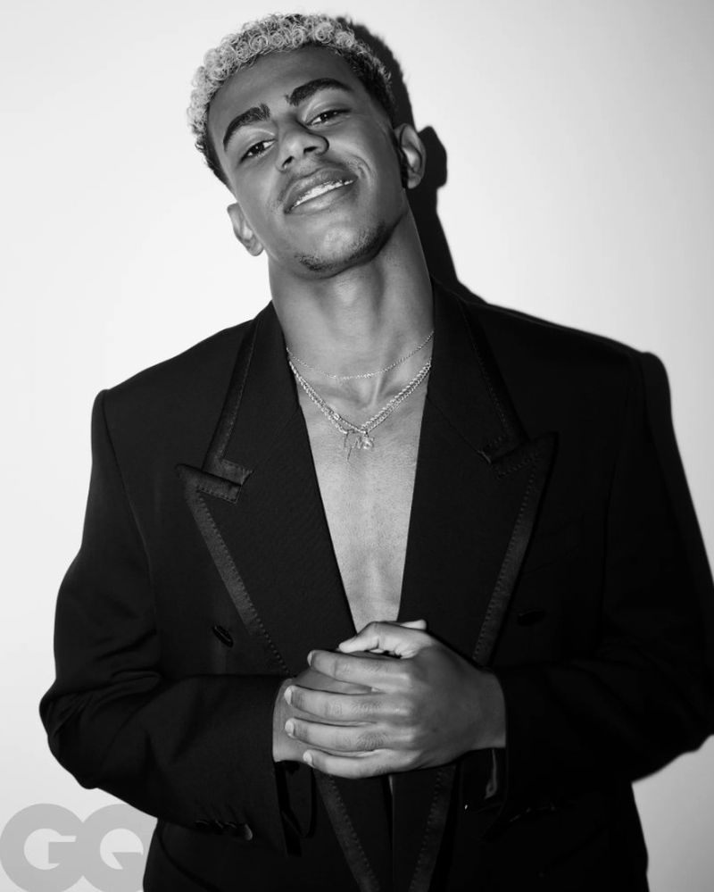 Monochrome portrait of a smiling light-skinned adult male with blonde hair wearing a black blazer and a silver necklace against a white wall.