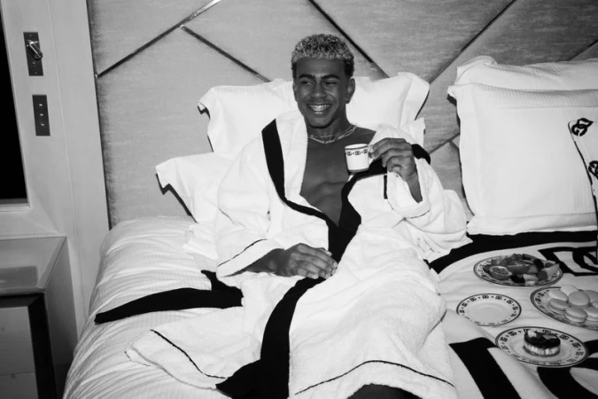 Smiling man in bathrobe sits in bed holding a teacup with plates of fruit and macarons in a monochrome indoor setting.