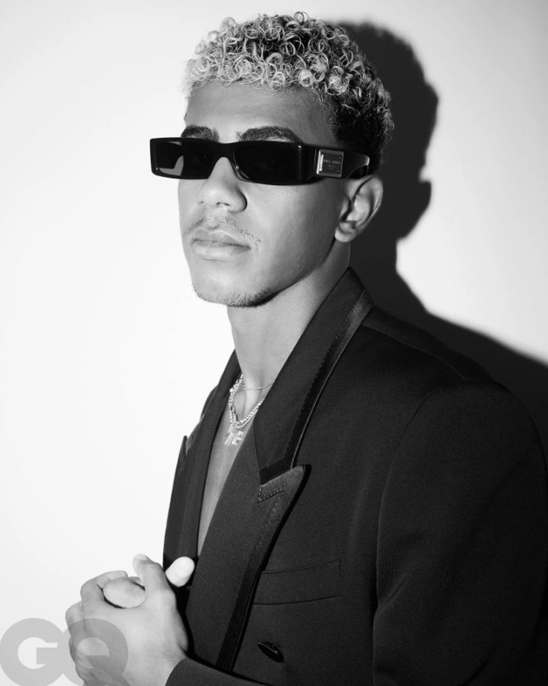 Black and white portrait of a young man with dark sunglasses wearing a black blazer and a silver chain against a gradient background.