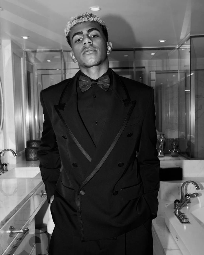 Black and white portrait of a young adult man in a formal black suit with a bow tie, posing in a modern bathroom with reflective surfaces.