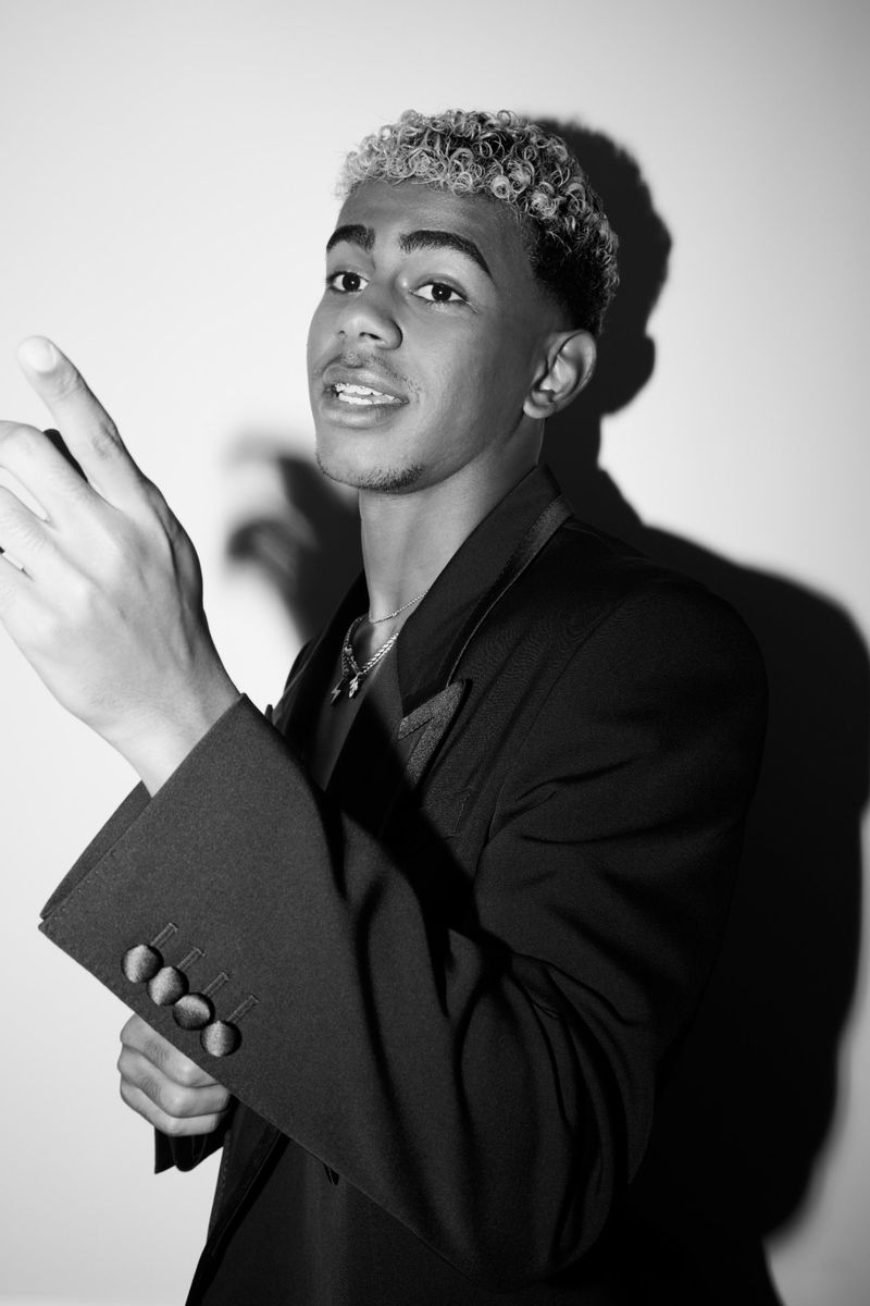 Black and white portrait of a young adult male with short curly hair, wearing a blazer, and pointing to the upper-left against a white background.