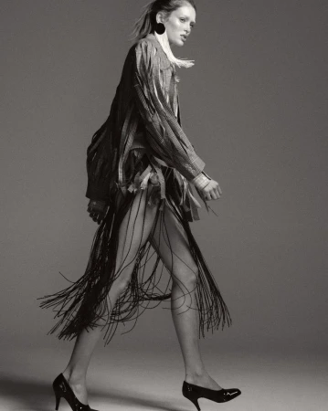 Model wearing fringed skirt and textured jacket walking against a plain grey studio backdrop