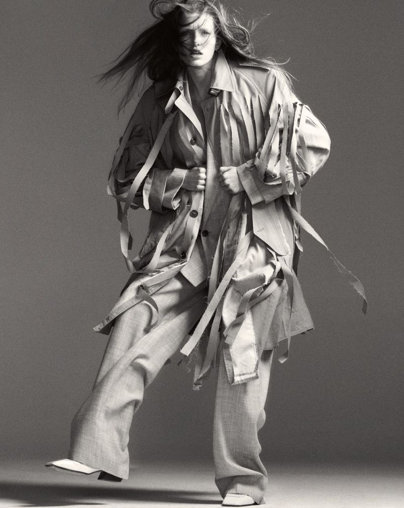 Monochromatic fashion shot: an adult female strides forward, wearing a deconstructed trench coat and wide-leg pants against a grey backdrop.