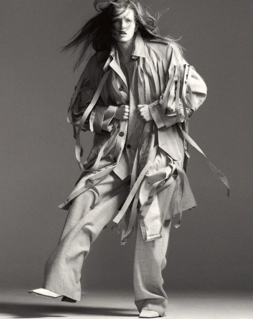 Monochromatic fashion shot: an adult female strides forward, wearing a deconstructed trench coat and wide-leg pants against a grey backdrop.