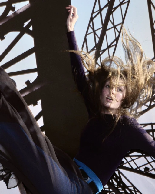 Woman with long hair in a black shirt and blue pants posing against a metallic tower structure with a light blue sky backdrop.