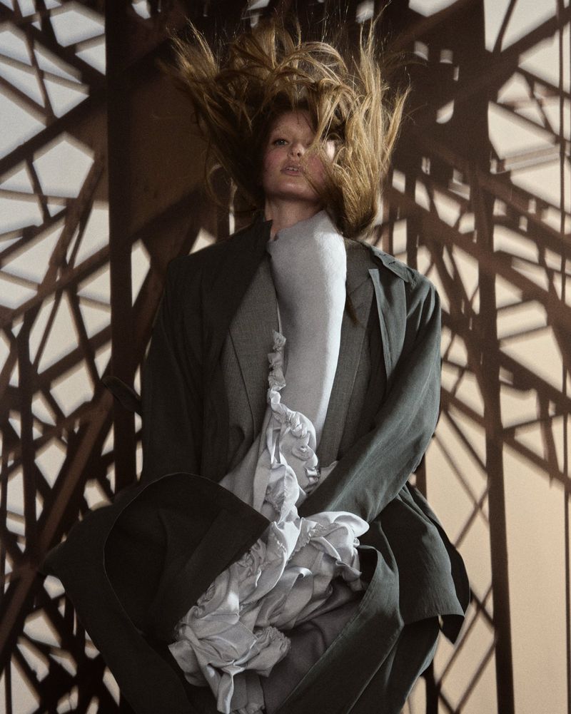 Fashionable woman in a grey coat poses in front of the Eiffel Tower structure with windblown hair.