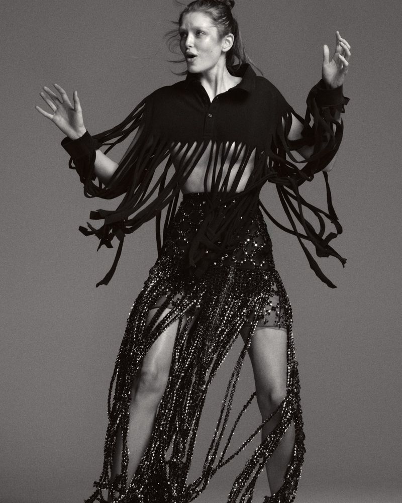 Adult woman in a black fringed top and skirt with sequins stands in a photo studio. Arms raised with a neutral facial expression.