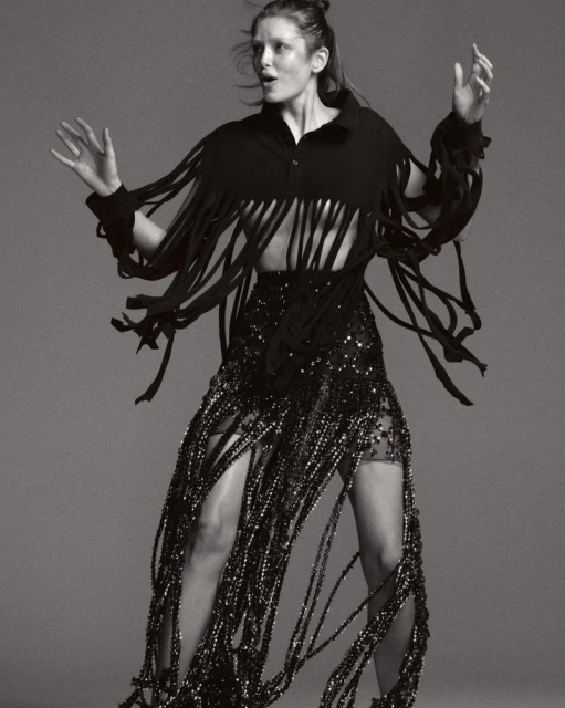 Adult woman in a black fringed top and skirt with sequins stands in a photo studio. Arms raised with a neutral facial expression.